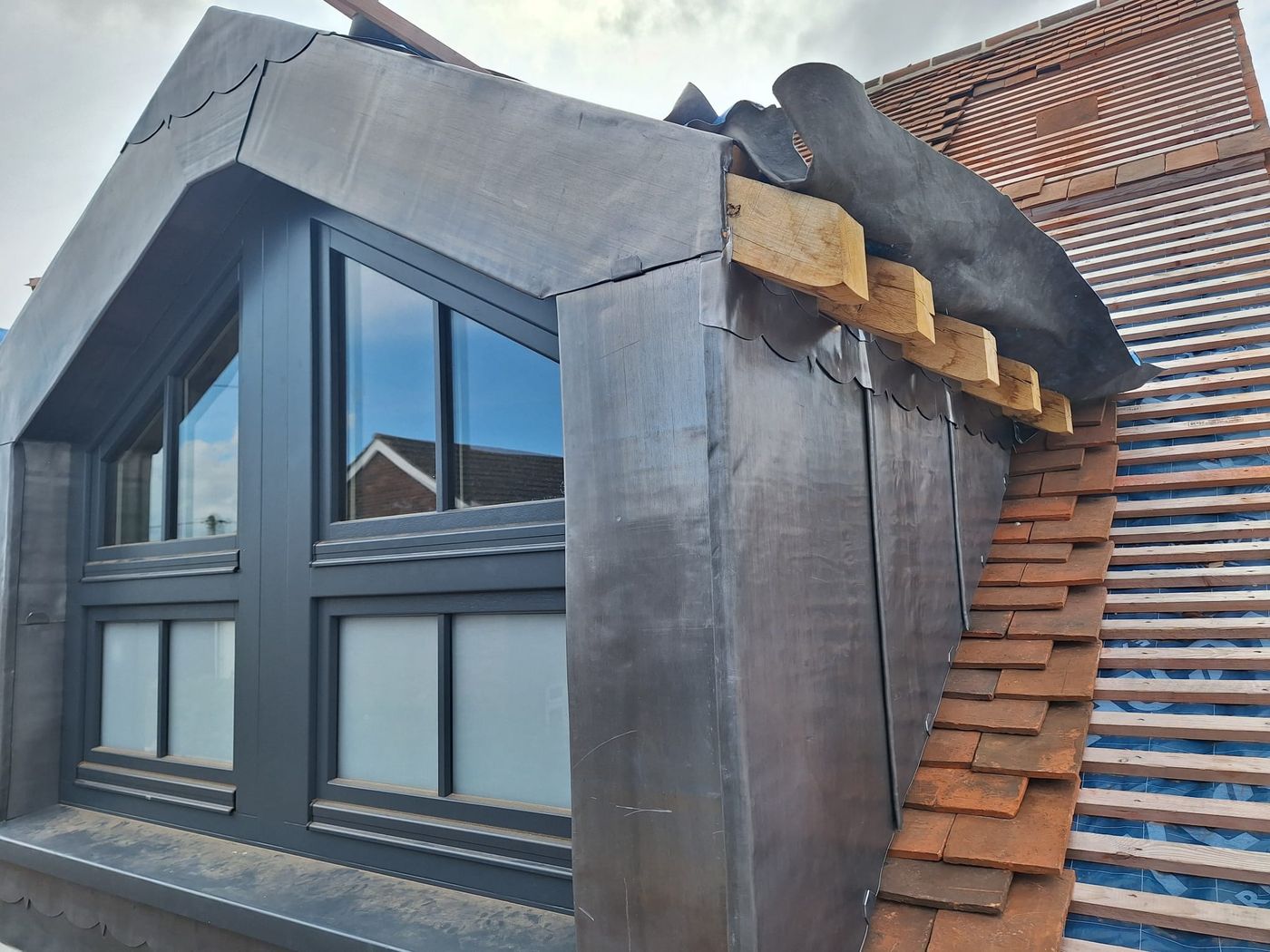 Side cheek of a lead-clad dormer with scalloped lead trim, oak timbers and new clay tiles being laid on battens
