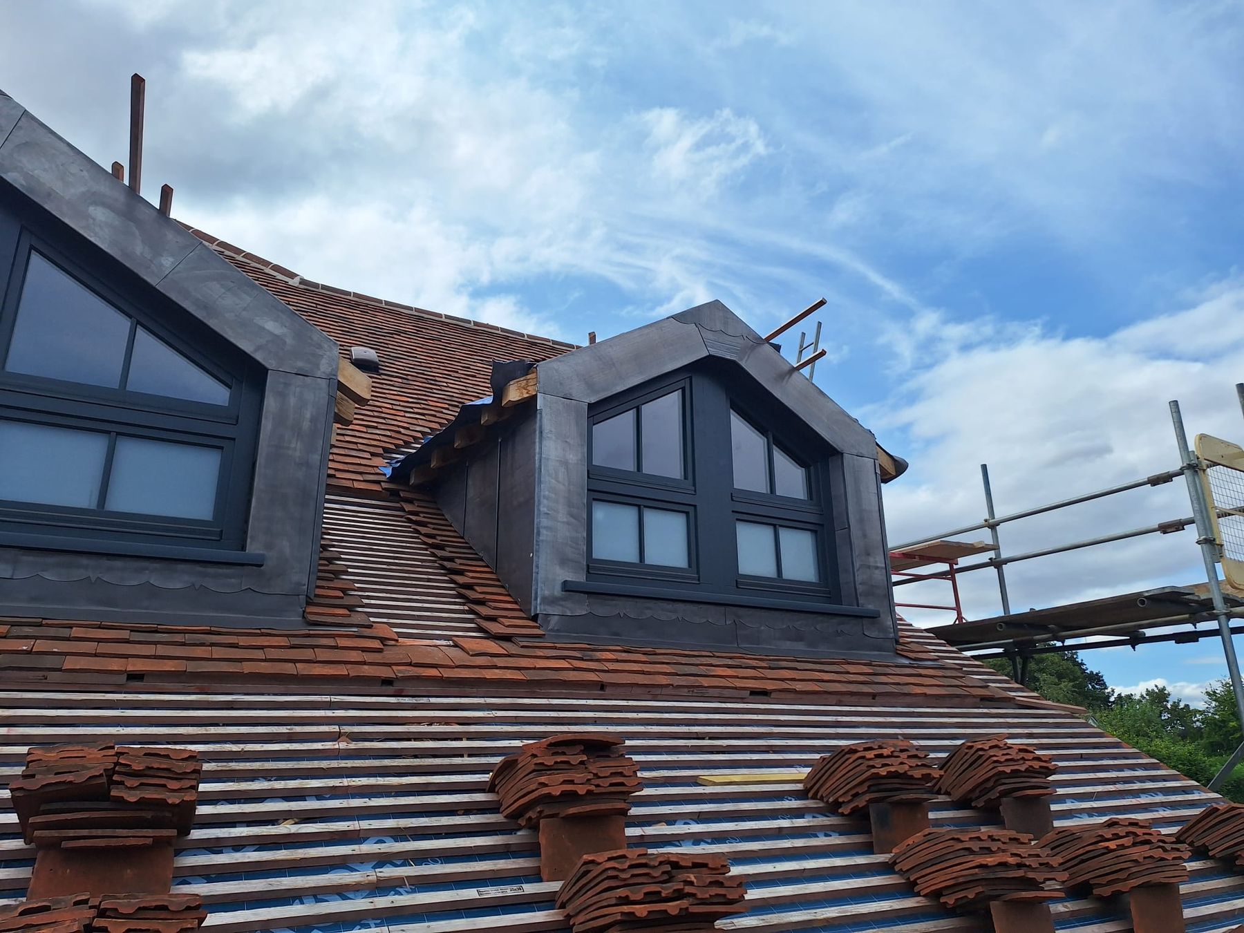 Plain clay tile roof being laid around two lead-clad dormers, with tiles stacked on the battens and scaffolding alongside
